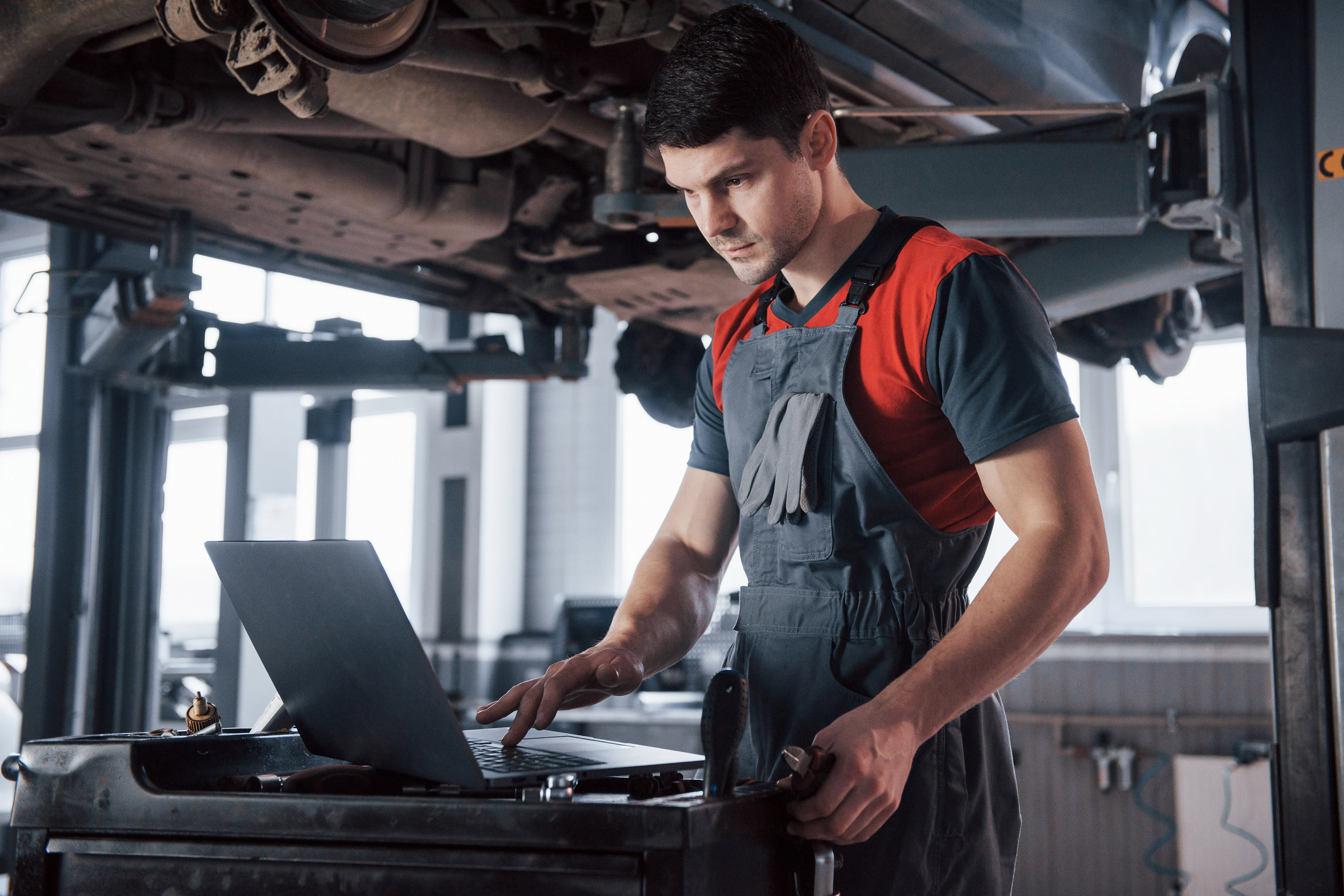 Silver colored device. Man at the workshop in uniform using laptop for his job for fixing broken car
